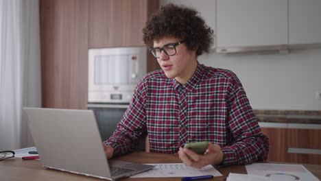 Young-man-with-smartphone-in-his-hands.-Modern-businessman-or-student-at-home-office.-Freelancer-at-work.-Young-student-man-study-at-home-with-laptop-and-uses-a-smartphone